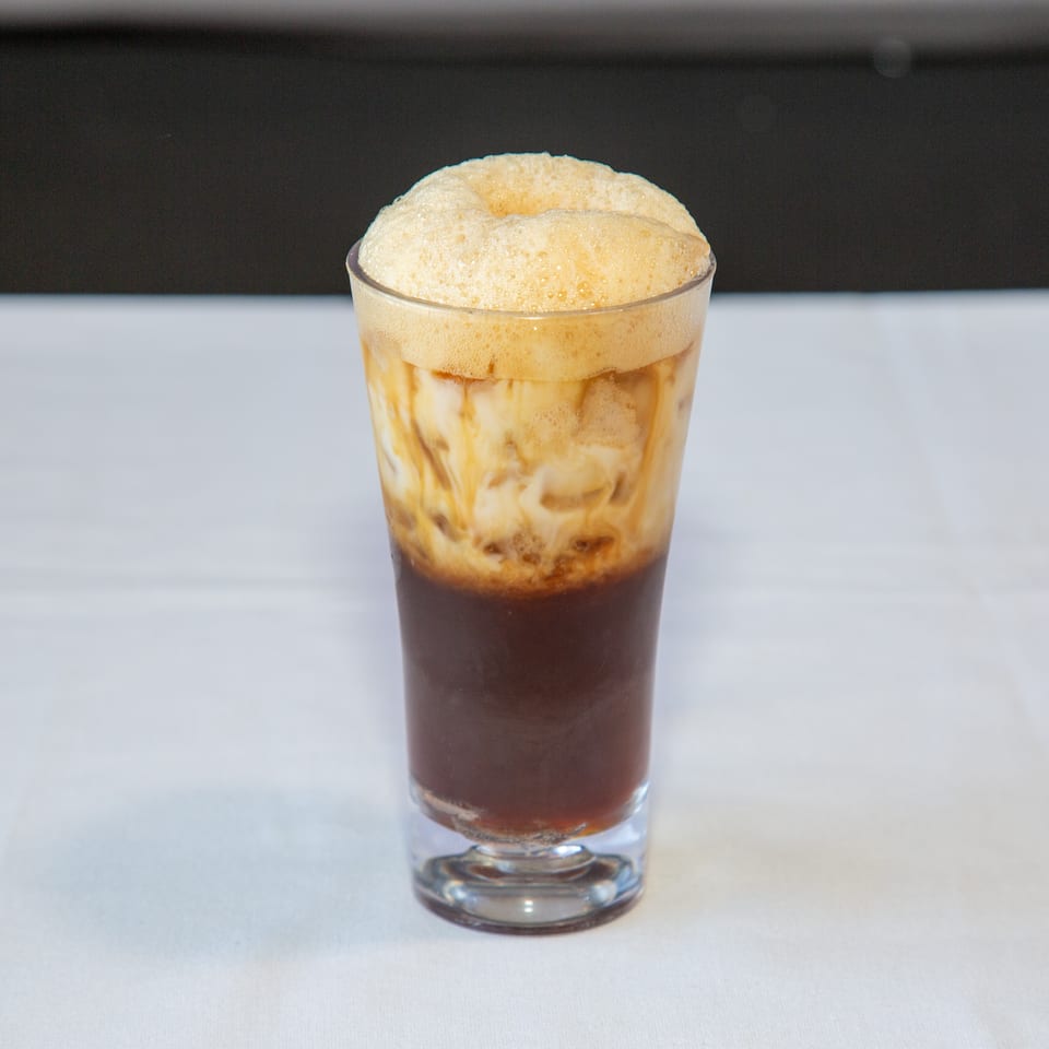 Tall glass with a dark beverage, creamy milk, and a thick layer of foam.