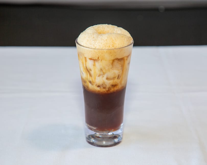 Tall glass with a dark beverage, creamy milk, and a thick layer of foam.