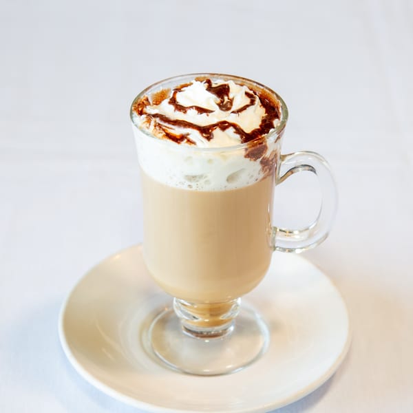 Glass mug of coffee with whipped cream and chocolate drizzle on a white saucer.