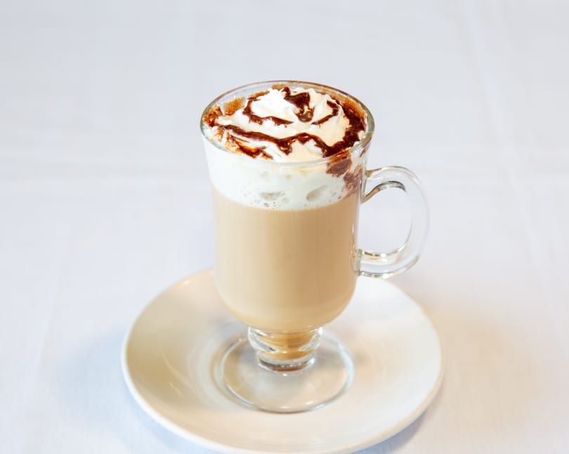 Glass mug of coffee with whipped cream and chocolate drizzle on a white saucer.