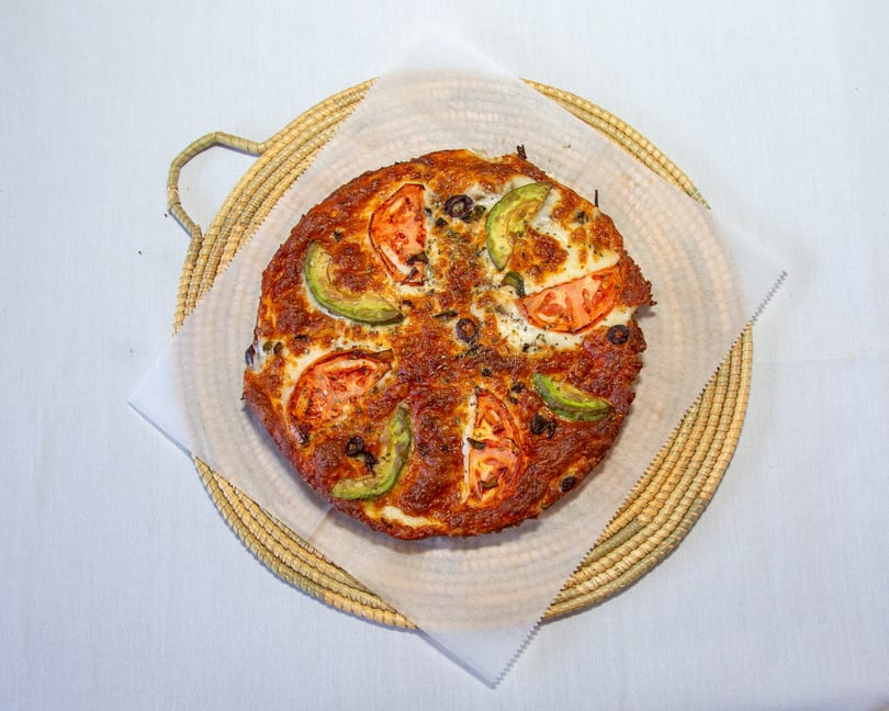 Baked pizza topped with melted cheese, tomato slices, avocado, and olives on a woven tray.