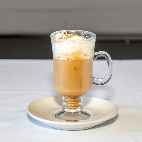 Latte with whipped cream and cinnamon in a clear glass mug on a white saucer.