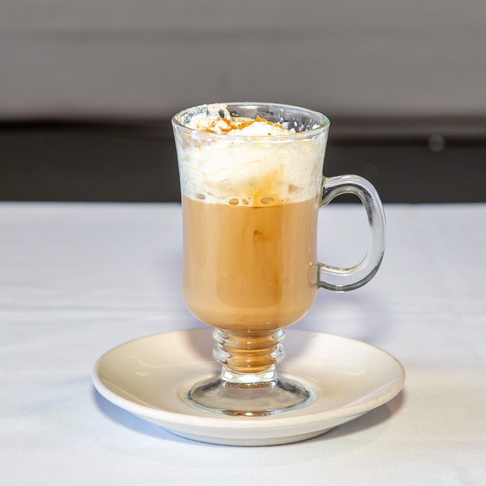 Latte with whipped cream and cinnamon in a clear glass mug on a white saucer.