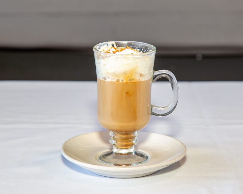 Latte with whipped cream and cinnamon in a clear glass mug on a white saucer.