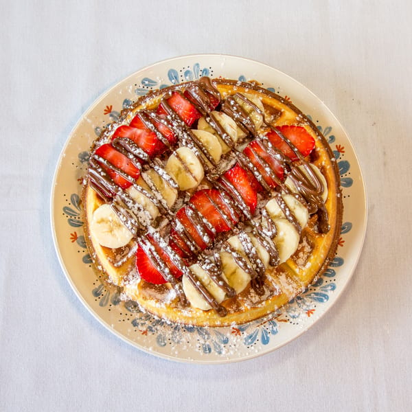 Round waffle topped with sliced strawberries, bananas, chocolate drizzle, and powdered sugar on a decorative plate.