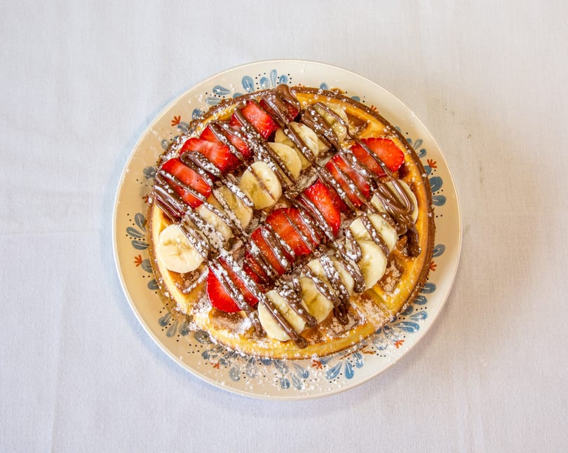 Round waffle topped with sliced strawberries, bananas, chocolate drizzle, and powdered sugar on a decorative plate.